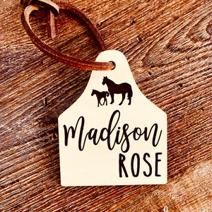 May include: A white wooden horse tag with a brown leather strap. The tag features a silhouette of a horse and foal with the name "Madison ROSE" printed in black.