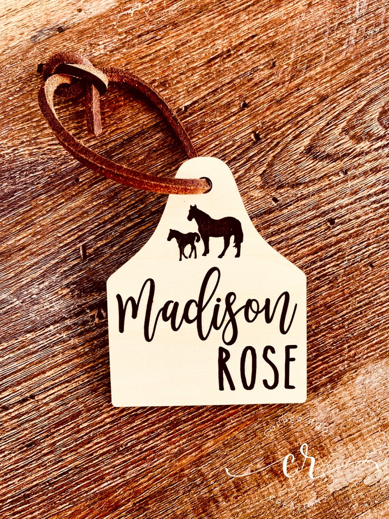 May include: A white wooden horse tag with a brown leather strap. The tag features a silhouette of a horse and foal with the name "Madison ROSE" printed in black.