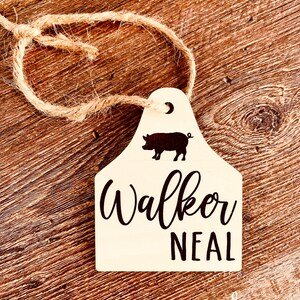 May include: A white wooden tag with a black pig silhouette, a crescent moon, and the text "Walker Neal". The tag is attached to a piece of twine.