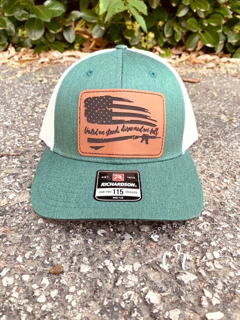 May include: A green and white trucker hat with a brown leather patch featuring a distressed American flag and the text "United we stand disarmed we fall". The patch also includes a silhouette of a rifle. The hat has a Richardson brand label.