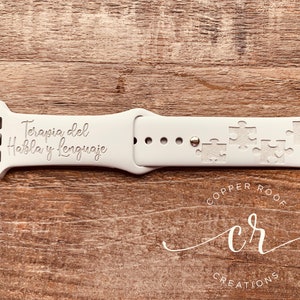 May include: White Apple Watch band with the text "Terapia del Habla y Lenguaje" engraved on it. The band also features a design of puzzle pieces. The band is made by Copper Roof Creations.
