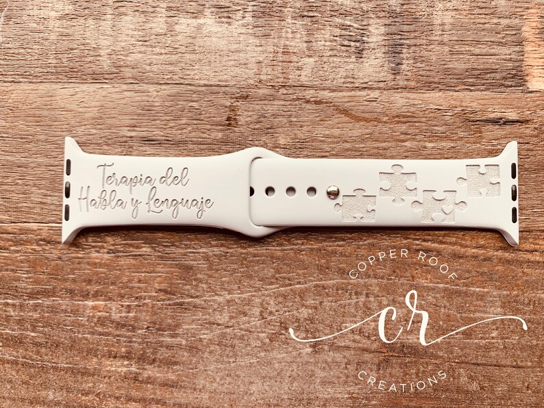 May include: White Apple Watch band with the text "Terapia del Habla y Lenguaje" engraved on it. The band also features a design of puzzle pieces. The band is made by Copper Roof Creations.