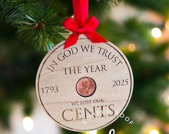 May include: A round wooden Christmas ornament with a penny in the center, hanging from a red ribbon. The ornament has the text "IN GOD WE TRUST", "THE YEAR", "WE LOST OUR CENTS", and the years 1793 and 2025.