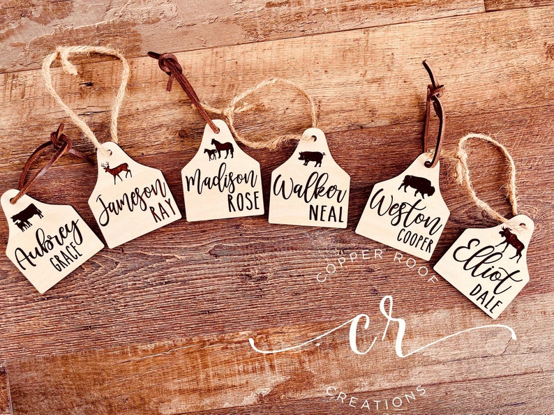 May include: Six wooden personalized tags shaped like cattle ear tags with a leather cord. Each tag features a different animal silhouette and a name. The names are Aubrey Grace, Jameson Ray, Madison Rose, Walker Neal, Weston Cooper, and Elliot Dale.