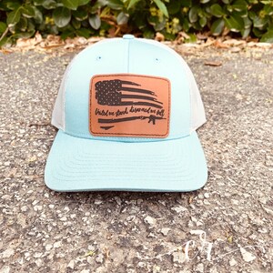 May include: A light blue and white trucker hat with a brown leather patch featuring a distressed American flag and the text "United we stand, disarmed we fall".