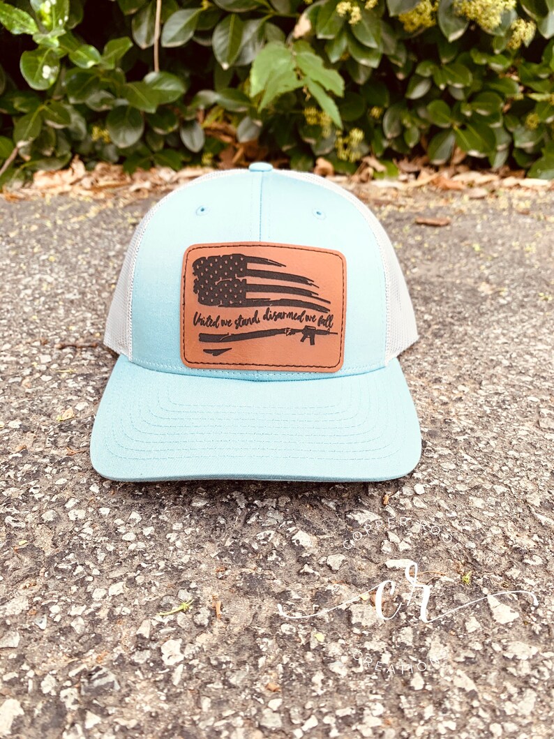 May include: A light blue and white trucker hat with a brown leather patch featuring a distressed American flag and the text "United we stand, disarmed we fall".