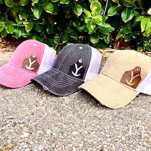 May include: Three trucker hats with a white mesh back and a brown leather patch with a white anchor design. The hats are in pink, black, and beige.