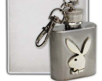 May include: A silver-colored flask with a screw-on cap and a hinged clasp. The flask features the Playboy bunny logo on the front. A rectangular, white object is behind the flask.