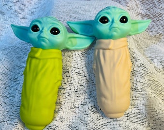 May include: Two Grogu-themed bottle stoppers. The stoppers feature Grogu's head in a light teal color with large black eyes and pointed ears. One stopper has a lime green body, and the other has a tan body. The stoppers are on a white, textured surface.