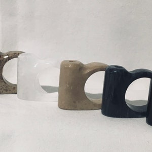 May include: Five different colored stone rings, each with a unique marbled pattern. The rings are arranged in a row from left to right, starting with pink, then brown, white, beige, and black.