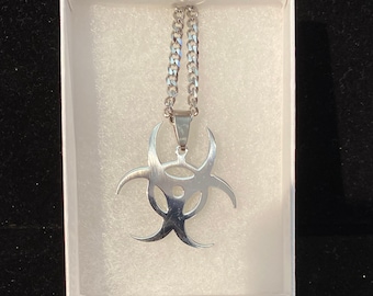 May include: A silver necklace with a biohazard symbol pendant. The necklace is in a white box with white foam padding.