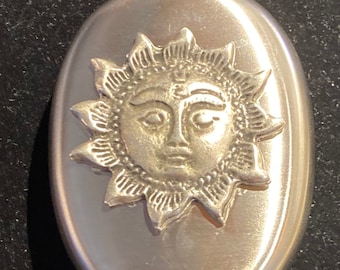 May include: A silver oval-shaped flask with a sun face design. The sun has a face with eyes, a nose, and a mouth. The sun is surrounded by rays.