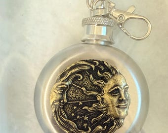 May include: A silver-colored flask with a decorative gold-toned face of the sun and moon. The flask has a silver cap and a silver clip for attaching to a belt loop or bag. The flask is likely for carrying liquor.