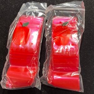 May include: Two packages of red plastic zipper bags with an apple logo and the text "TM" on each bag. The bags are stacked and sealed in clear plastic.