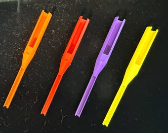 May include: Four elongated plastic tools in bright orange, red, purple, and yellow. Each tool features a wide, U-shaped channel at the top that tapers into a narrower, straight handle section with a recessed groove running along the center.