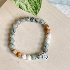 May include: A bracelet made of round beads in shades of light green, brown, and white. It has a silver charm with a tree design. The beads are strung on an elastic band.