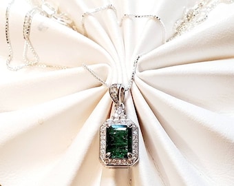 May include: A silver necklace with a green emerald pendant surrounded by smaller diamonds. The necklace is on a white fabric background.