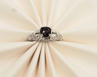 May include: A silver ring with a dark purple gemstone surrounded by smaller clear gemstones. The ring is on a white background.