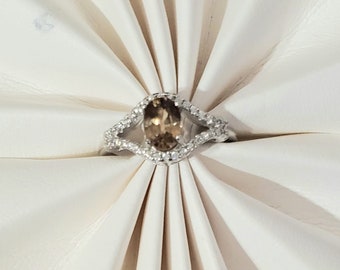 May include: A silver ring with an oval, light brown gemstone at the center, surrounded by smaller, clear stones. The ring is set on a white, folded surface, creating a layered effect. The ring's design features a split band with additional small stones.