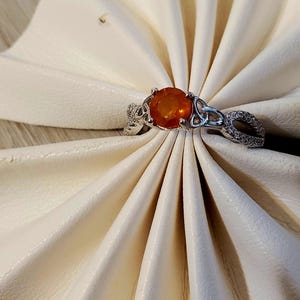 May include: A silver ring with an oval, orange gemstone. The ring features a Celtic knot design on the band and around the gemstone. The ring is displayed on a white, textured surface.