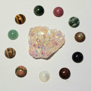 May include: A collection of 11 round, polished gemstone cabochons in various colors and patterns, including green, brown, pink, black, and white. The stones are arranged in a circle around a large, white, iridescent crystal.