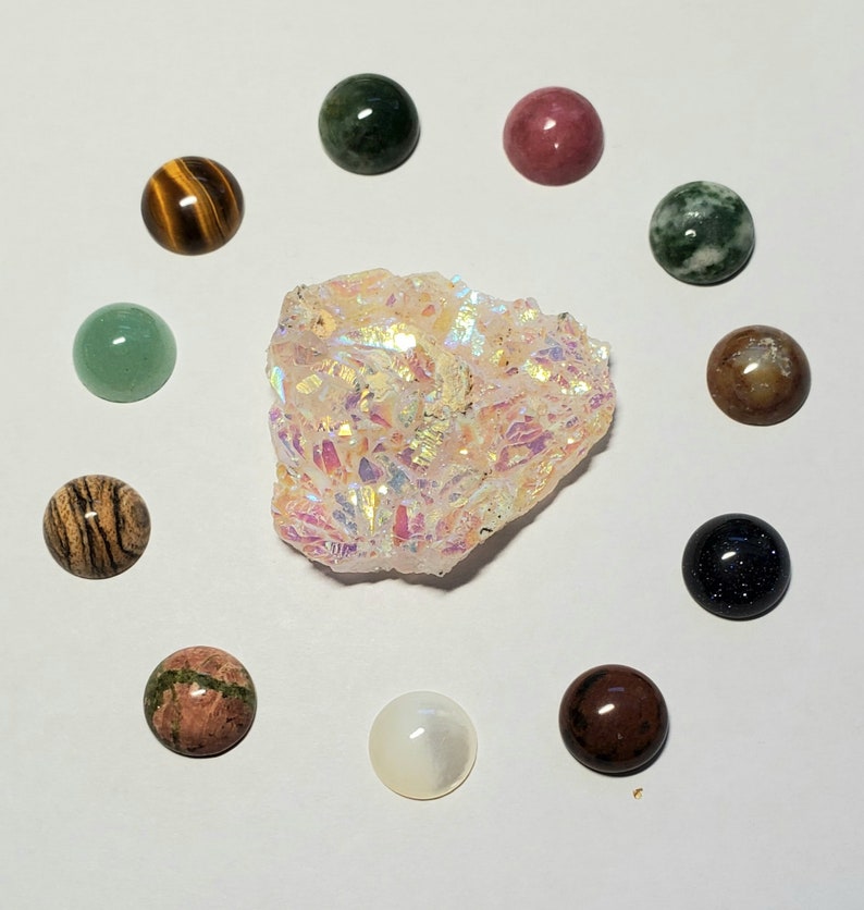 May include: A collection of 11 round, polished gemstone cabochons in various colors and patterns, including green, brown, pink, black, and white. The stones are arranged in a circle around a large, white, iridescent crystal.