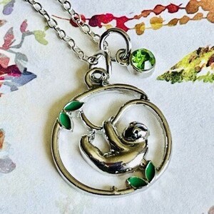 May include: Silver necklace with a green gemstone charm and a pendant featuring a sloth hanging from a branch with green leaves.