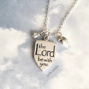 May include: Silver necklace with a shield-shaped pendant that reads "the Lord be with you." The pendant is attached to a silver chain with a small crystal charm and a heart charm.
