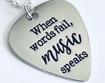 May include: Silver-toned guitar pick pendant necklace with a chain. The pendant has the words "When words fail, MUSIC speaks" engraved in black. The necklace is on a white background.