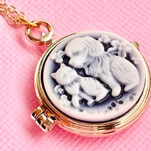 May include: Gold tone necklace with a round locket featuring a white cameo of a dog and cat. The cameo is set against a dark blue background.