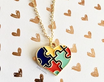 May include: A gold chain necklace with a heart-shaped pendant. The pendant is made of metal and is painted with a puzzle design in blue, yellow, red, and green. The background is a white surface with a pattern of brown hearts.