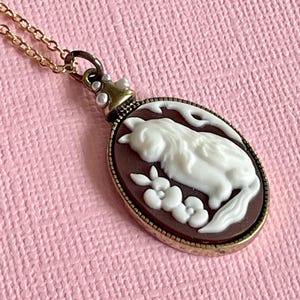 May include: A gold-toned necklace featuring an oval cameo pendant. The pendant showcases a white cat design on a brown background, with floral accents. The chain is delicate, and the pendant has a detailed, vintage aesthetic.