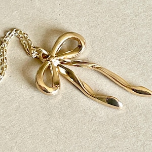 May include: A gold-colored bow pendant necklace. The bow is detailed with curved lines and a textured surface. The necklace chain is also gold and delicate. The pendant is a classic jewelry design.