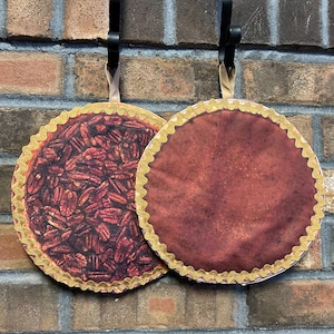 May include: Two decorative pie-shaped pot holders hanging on hooks. One pot holder has a pecan pie design, the other is a solid brown color. Both have a gold scalloped edge.