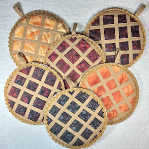 May include: Six quilted pot holders shaped like pie crusts. The pot holders have different fillings: orange, red, and blue. Each pot holder has a loop handle.