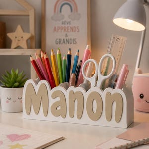 Peut inclure: Un organisateur de bureau blanc avec le prénom Manon en lettres beiges en relief sur le devant. L'organisateur contient divers crayons de couleur, une paire de ciseaux blancs, une règle en plastique transparent et des stylos pour le rangement.