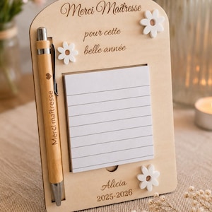 Peut inclure: Un organisateur de bureau en bois clair comprenant un bloc-notes vertical à feuilles blanches lignées, un stylo en bambou avec le texte "Merci maîtresse" et une icône de cœur, ainsi que trois décorations en forme de fleurs blanches. Le support est gravé "Merci Maîtresse pour cette belle année", "Alicia" et "2025-2026".