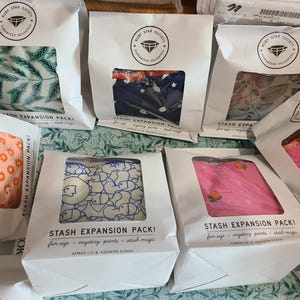 May include: Several white paper bags with clear windows displaying colorful fabric patterns. The bags are labeled "STASH EXPANSION PACK!" and feature the Ruby Star Society logo. The fabrics include floral, leaf, and geometric designs.