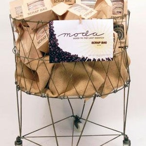 May include: A wire basket on wheels filled with brown paper bags labeled "Scrap Bag." A sign reading "moda GOOD TO THE LAST SWATCH SCRAP BAG" is attached. The basket has a metal frame and is lined with tan fabric.