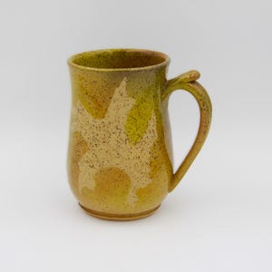 May include: A handmade ceramic mug with a unique design. The mug features a speckled, mottled pattern in shades of yellow and green, with a curved handle. The mug is suitable for hot or cold beverages.