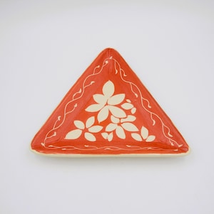 May include: A triangular ceramic dish with a vibrant orange background. The dish features a cream-colored floral design in the center and a delicate white vine-like pattern along the edges. The dish is a decorative piece.