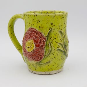 May include: A ceramic mug with a bright lime green glaze and dark brown speckles. The mug features a prominent, raised, three-dimensional red flower with a yellow center on the front, accompanied by sculpted green leaves extending along the side.