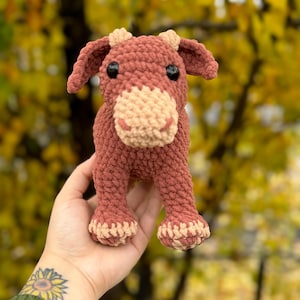 May include: A crocheted plush toy in the shape of a brown cow with a light brown nose and hooves. The toy is being held in a hand with a sunflower tattoo.
