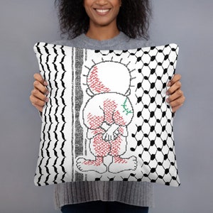 以下が含まれることがあります： 白地に黒と白の市松模様が両側に施されたクッション。クッションの中央には、腕を組んだ人物の赤と白のイラストが描かれている。人物の肩には緑色の植物が描かれている。