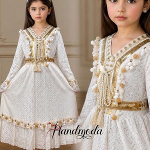 Può includere: Un vestito bianco con maniche lunghe, caratterizzato da intricati motivi in pizzo e abbellimenti dorati. L'abito ha uno scollo a V, una cintura decorativa e dettagli con nappe. L'abito è indossato da una bambina. Il marchio "Handmoda" è visibile.