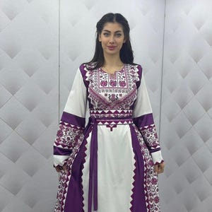 May include: A long, white dress with purple accents and floral embroidery. The dress features long sleeves, a cinched waist, and a decorative neckline. The dress has a flowing skirt and a matching purple belt.