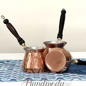 Puede incluir: Tres cafeteras turcas de cobre con mangos de madera. Las cafeteras tienen un acabado martillado y diseños florales intrincados. Las cafeteras están sobre una tela con un patrón azul y blanco.