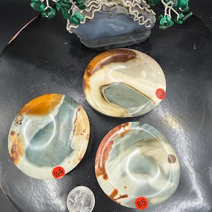 May include: Three polished stones with swirling patterns in shades of brown, cream, and blue-green. A quarter coin is next to the stones for size comparison. A wire tree sculpture with green stone leaves sits above the stones.