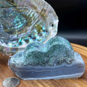 May include: A cloud-shaped, blue and green agate stone. The stone has a layered appearance with bands of blue and white. A quarter coin is next to the stone for size comparison. An iridescent abalone shell is in the background.