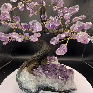 Könnte beinhalten: Eine handgefertigte Amethyst-Baumskulptur. Der Baum hat einen dunklen, strukturierten Stamm und Äste aus goldfarbenem Draht. Die Blätter sind polierte Amethyststeine. Die Basis ist ein Cluster aus Amethystkristallen, mit einer Münze als Größenreferenz.